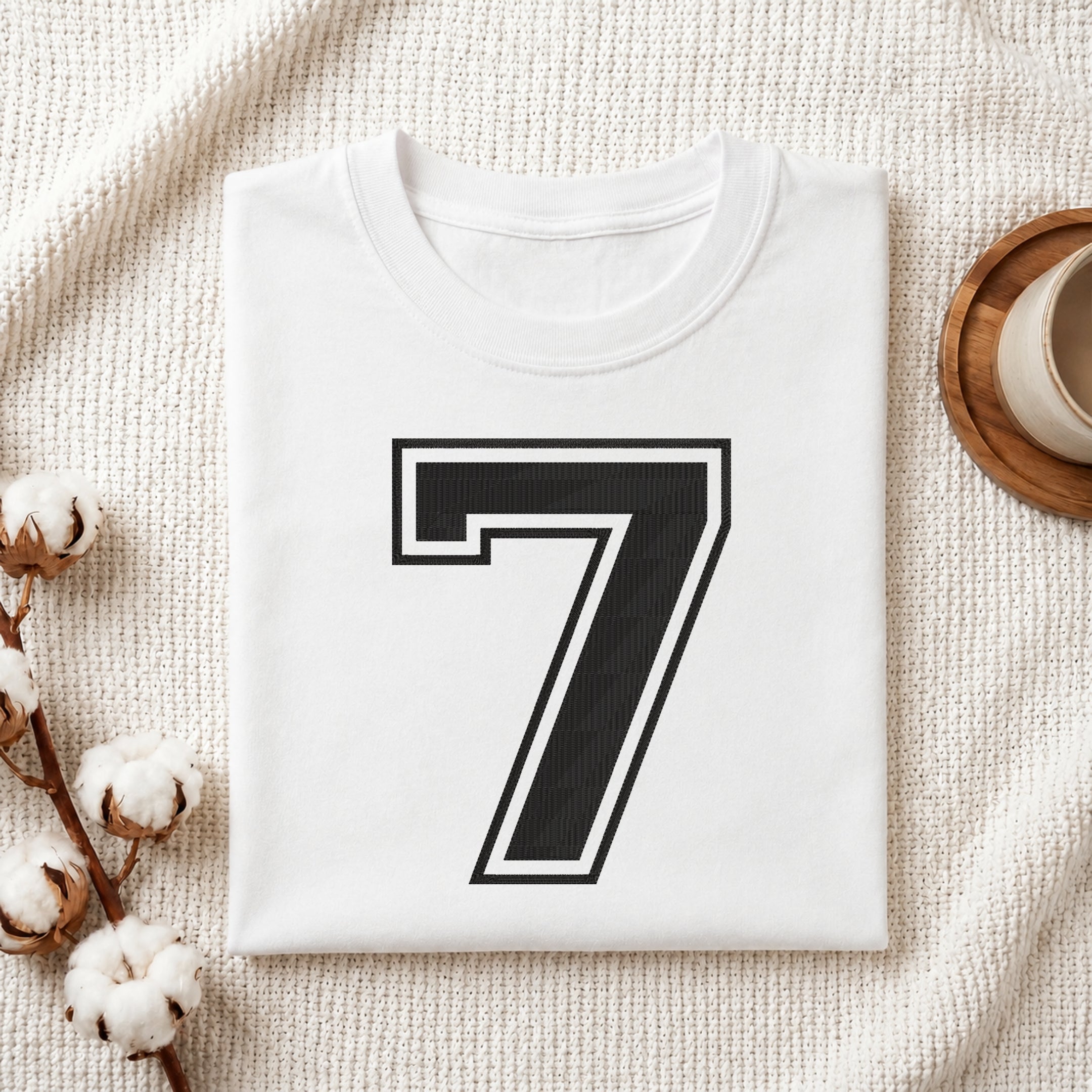A folded white T-shirt with a large black number 7 outlined in white on the front, styled on a textured cream fabric with cotton bolls and a small cup at the side.