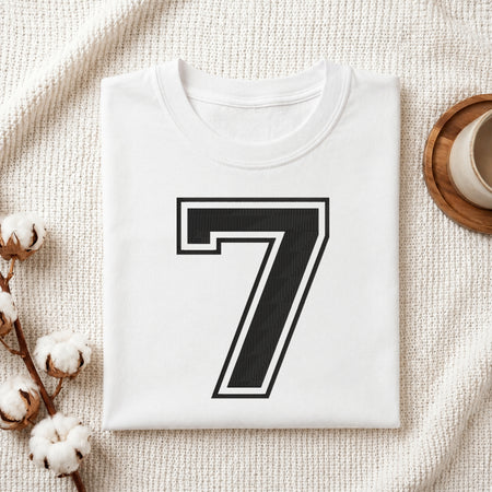 A folded white T-shirt with a large black number 7 outlined in white on the front, styled on a textured cream fabric with cotton bolls and a small cup at the side.