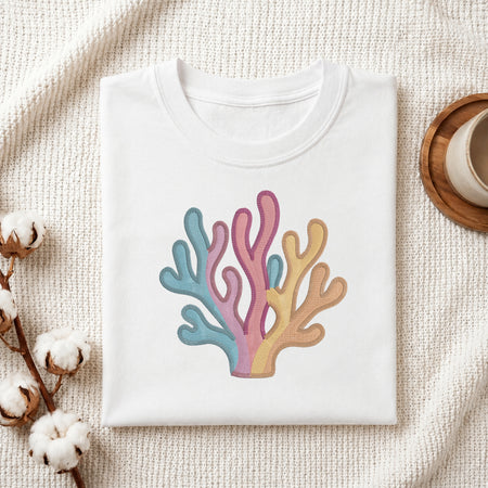 A folded white crewneck t-shirt featuring a colorful coral graphic in pastel blue, pink, and yellow-orange, styled on a textured cream knit blanket with cotton bolls and a wooden cup.