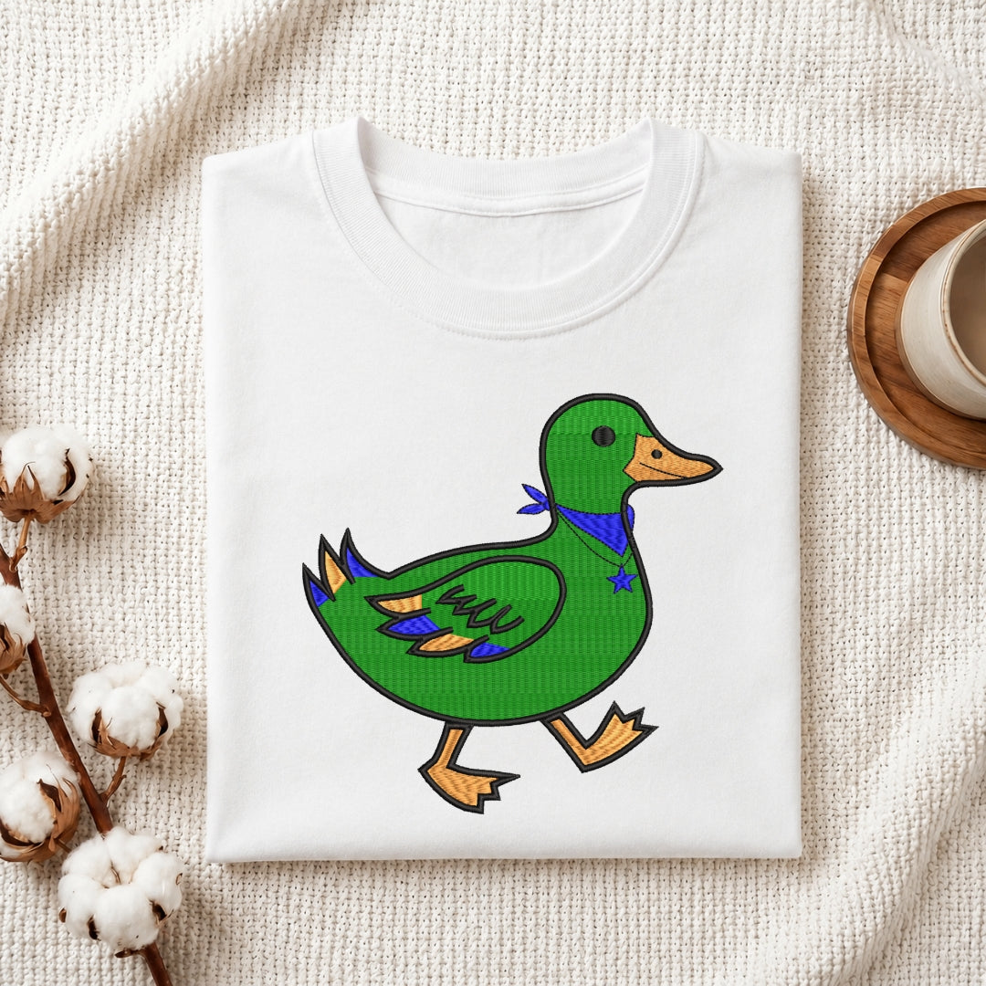 A folded white crewneck t-shirt featuring a cartoon illustration of a green duck wearing a blue bandana, styled on a textured cream fabric with cotton bolls and a wooden cup.