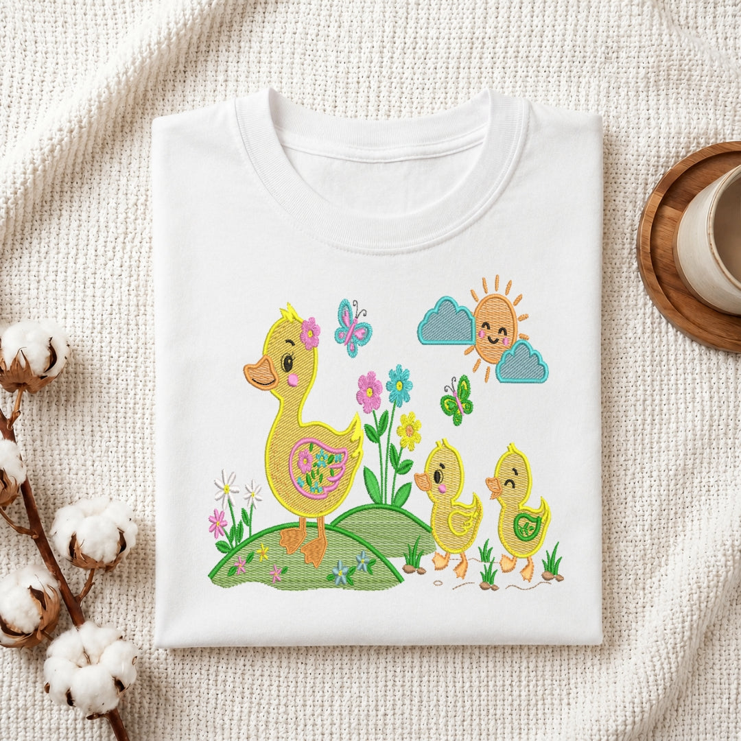 A folded white crewneck t-shirt with a colorful embroidered design of a mother duck, two baby ducks, flowers, butterflies, and a smiling sun, displayed on a textured cream fabric background with decorative cotton stems and a cup.