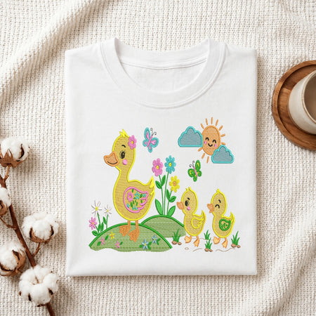 A folded white crewneck t-shirt with a colorful embroidered design of a mother duck, two baby ducks, flowers, butterflies, and a smiling sun, displayed on a textured cream fabric background with decorative cotton stems and a cup.