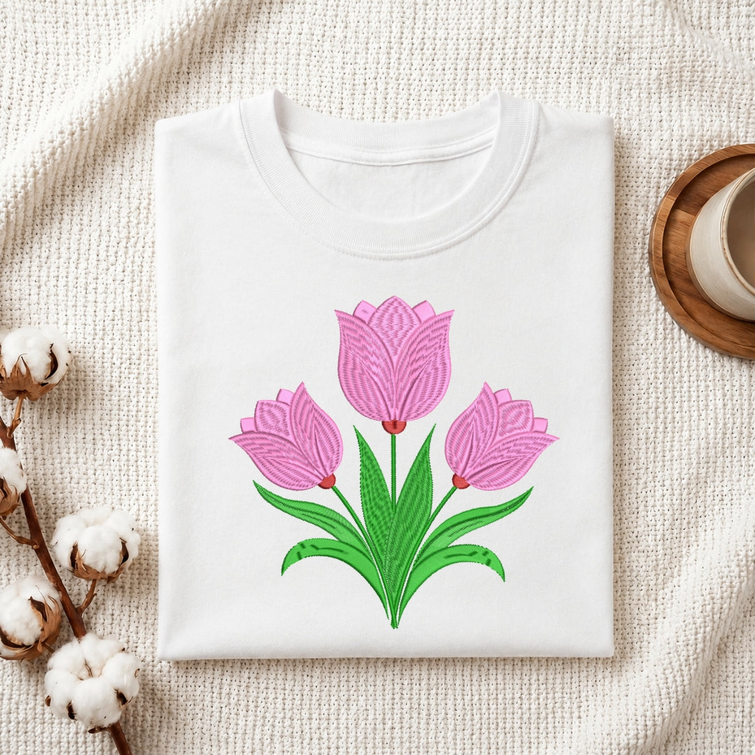 A folded white crewneck t-shirt featuring a graphic of three pink tulips with green leaves, styled on a textured cream fabric with cotton bolls and a wooden cup in the background.