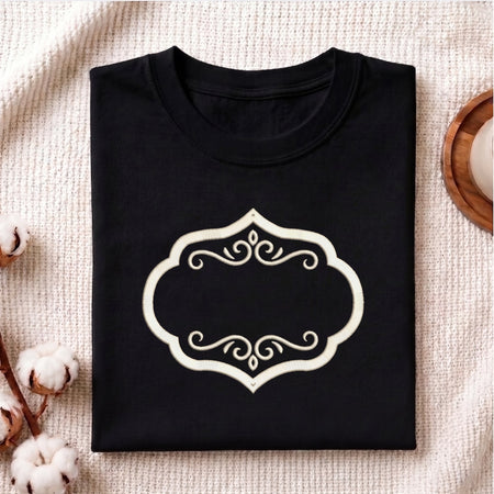 A folded black crewneck t-shirt with a cream-colored ornate frame graphic on the front, displayed on a textured beige fabric background with cotton bolls and a wooden slice prop.