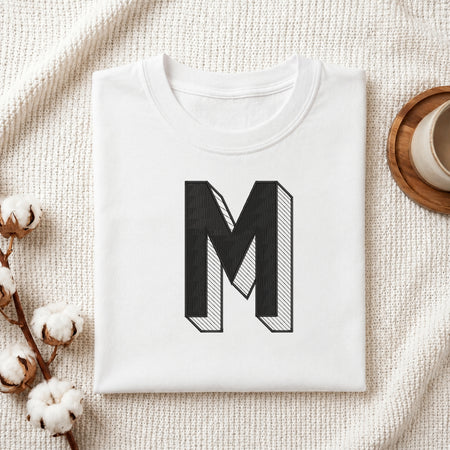 A folded white t-shirt with a large black block letter 'M' printed on the front, styled on a textured cream knit blanket with cotton bolls and a cup of coffee in the background.