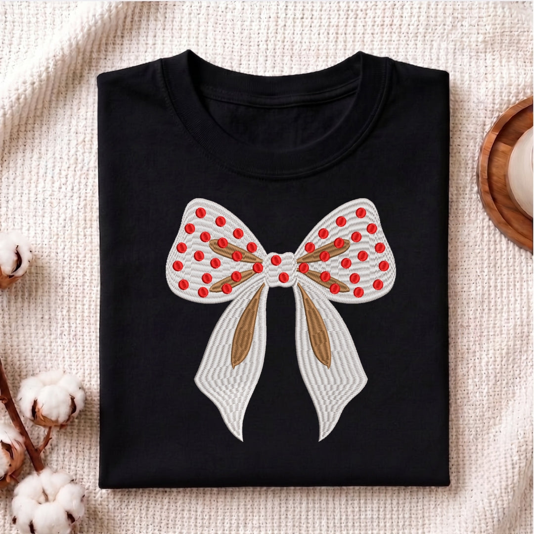 A folded black crewneck t-shirt featuring a large embroidered bow design on the front. The bow is white with red polka dots, a brown center, and white striped tails.