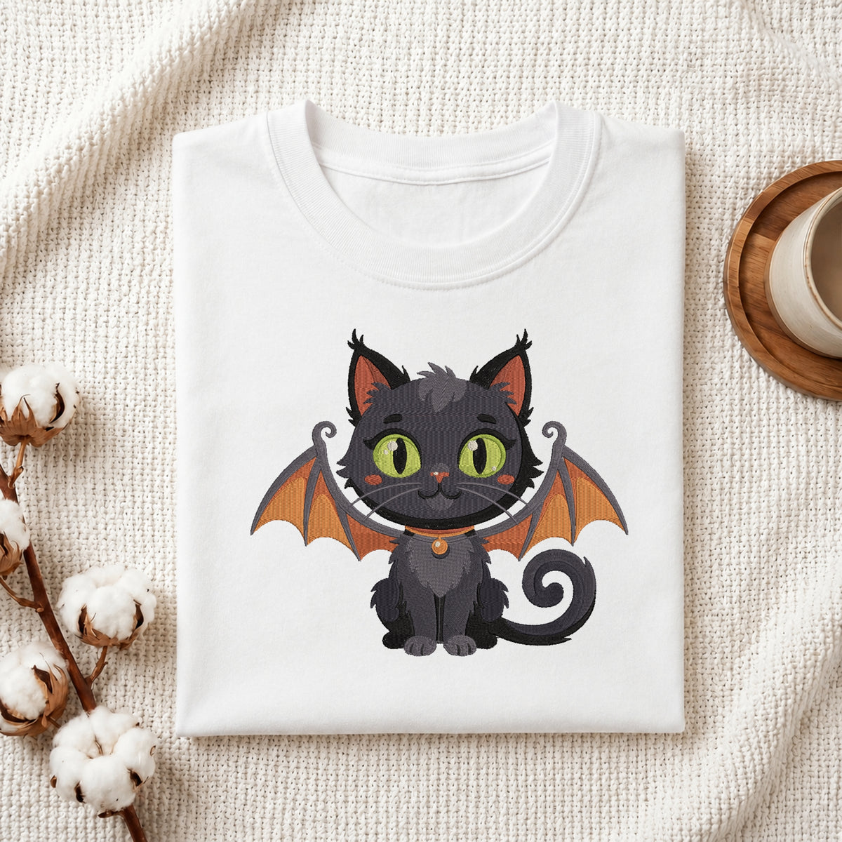 A neatly folded white crewneck t-shirt featuring a stitched Halloween bat cat design, styled with cotton branches and a wooden coaster.