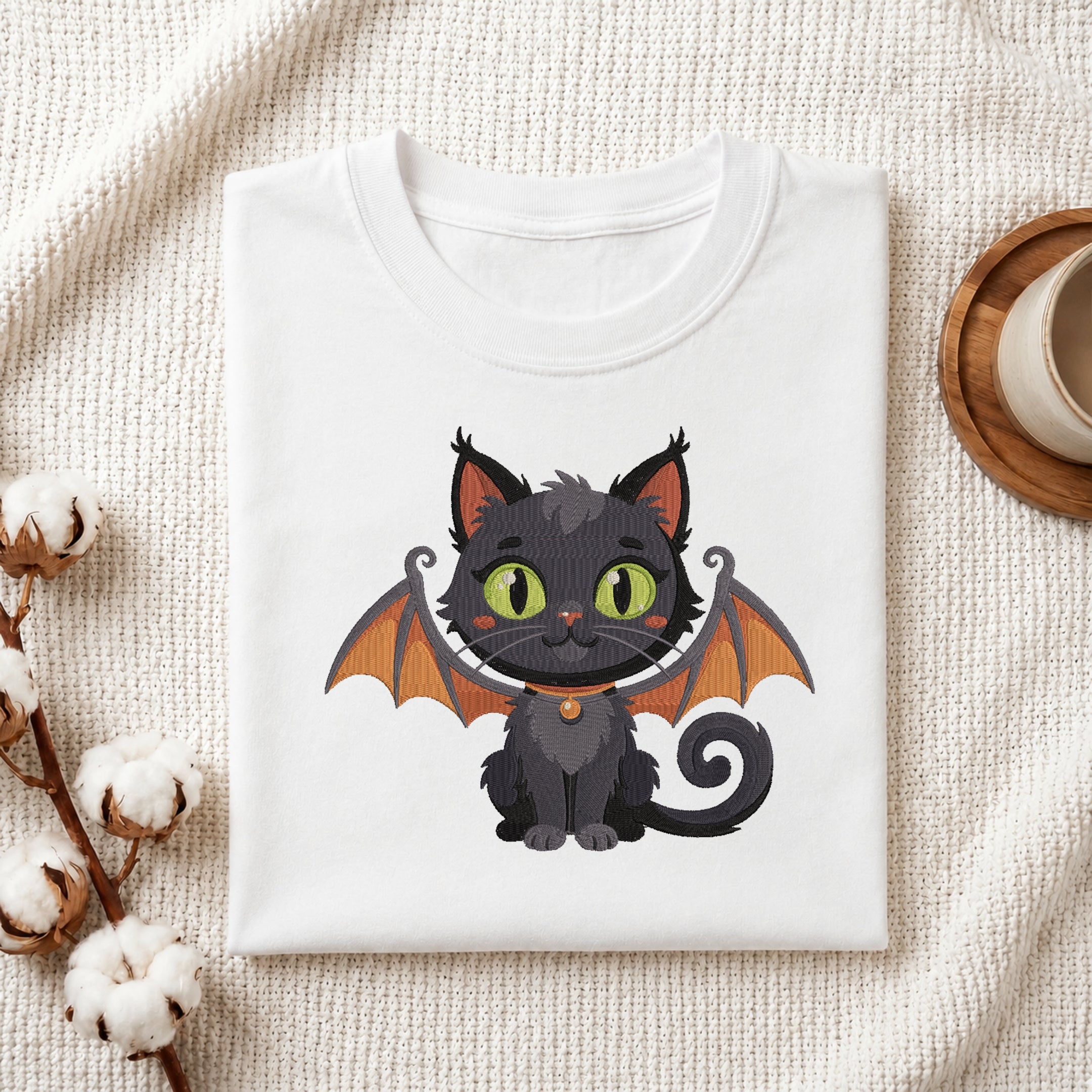A neatly folded white crewneck t-shirt featuring a stitched Halloween bat cat design, styled with cotton branches and a wooden coaster.