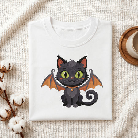 A neatly folded white crewneck t-shirt featuring a stitched Halloween bat cat design, styled with cotton branches and a wooden coaster.