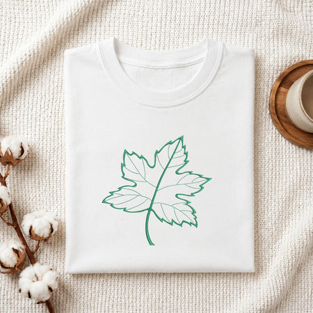 Folded white cotton t-shirt mockup featuring a green maple leaf machine embroidery design, styled with cotton stems and a wooden cup