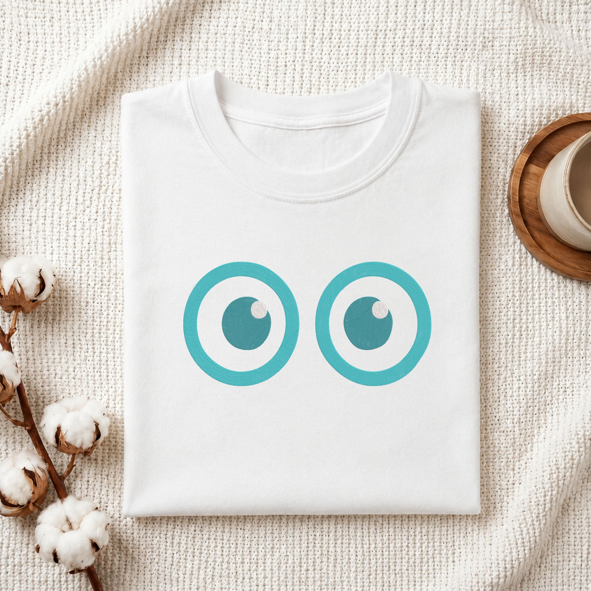 A folded white crewneck t-shirt with a graphic of two turquoise cartoon eyes, styled on a textured cream knit blanket with cotton bolls and a wooden cup in the background.