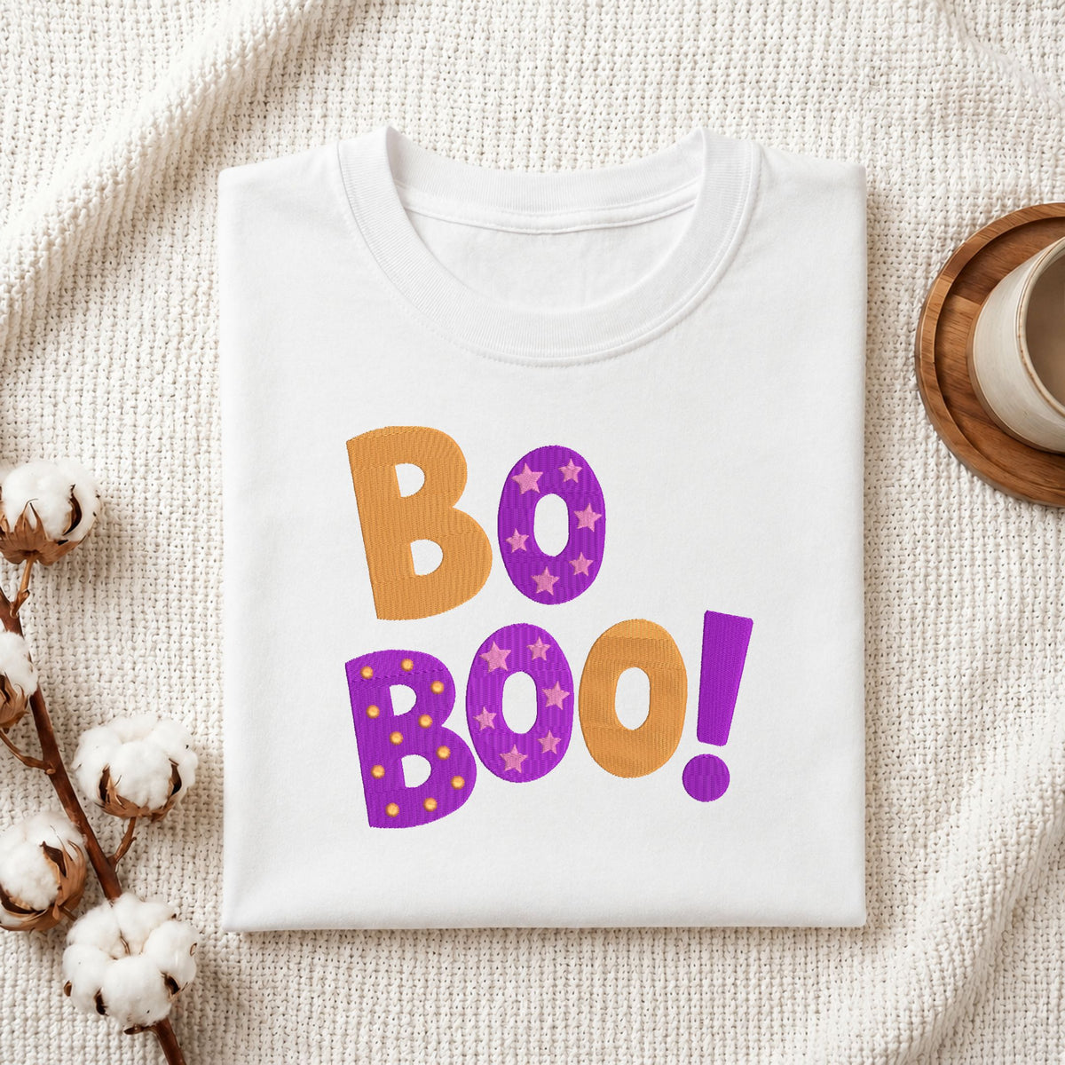 Folded white cotton t-shirt mockup featuring an orange and purple "Bo Boo!" Halloween machine embroidery design, styled with cotton stems and a wooden cup.