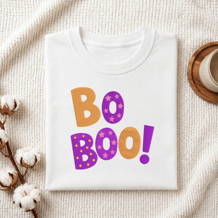 Folded white cotton t-shirt mockup featuring an orange and purple "Bo Boo!" Halloween machine embroidery design, styled with cotton stems and a wooden cup.