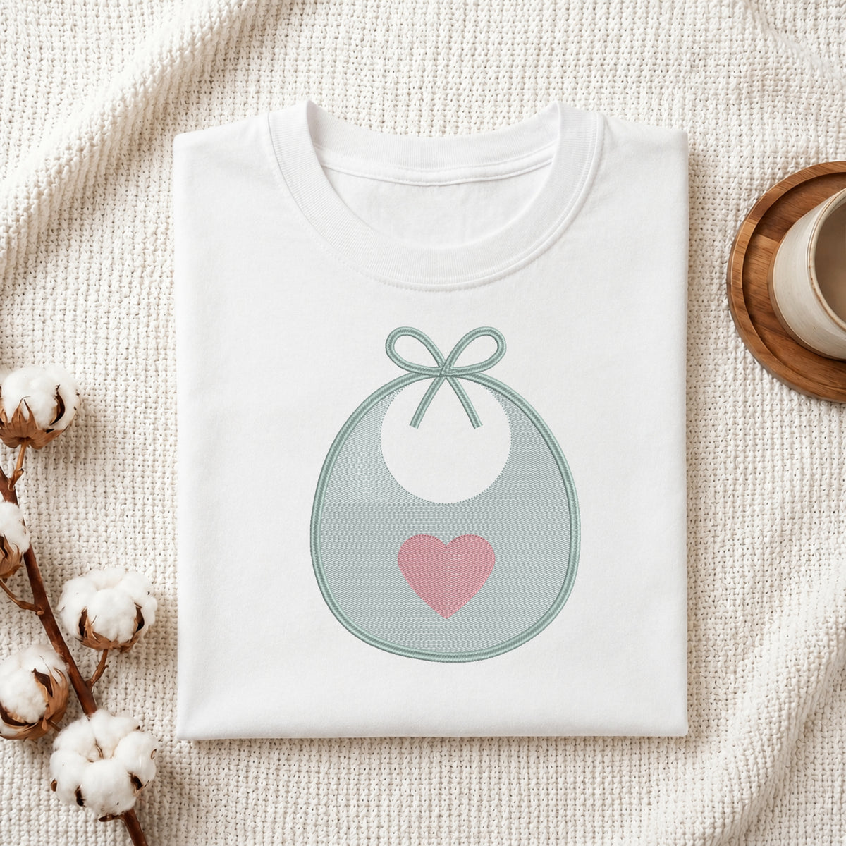 A folded white t-shirt with a graphic of a pastel blue-green bib with a pink heart, styled on a textured cream blanket with cotton bolls and a wooden cup.
