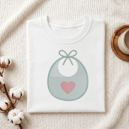 A folded white t-shirt with a graphic of a pastel blue-green bib with a pink heart, styled on a textured cream blanket with cotton bolls and a wooden cup.
