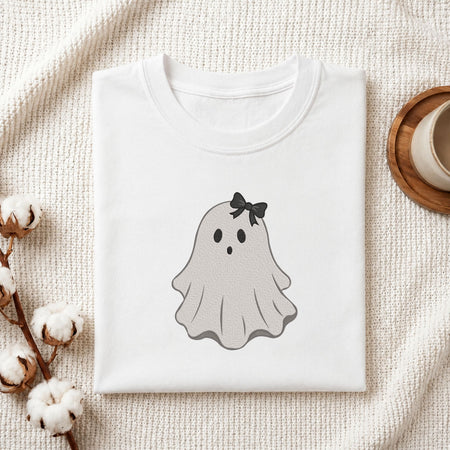 Folded white cotton t-shirt mockup featuring a cute ghost machine embroidery design, styled with cotton stems and a wooden cup