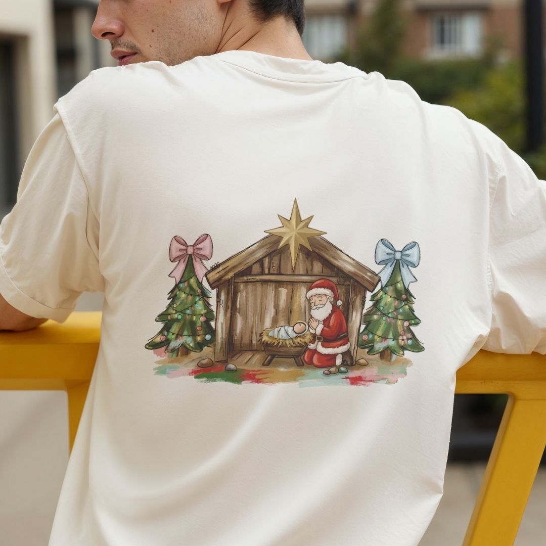A man wearing a cream-colored t-shirt, viewed from behind, which features a large watercolor-style graphic of a Christmas nativity scene on the back. The design includes a wooden stable with a golden star, Santa Claus, a baby in a manger, and two decorated Christmas trees with pink and blue bows.