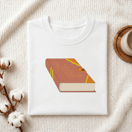 Folded white cotton t-shirt mockup featuring a magic spell book embroidery design, styled with cotton stems and a wooden cup.