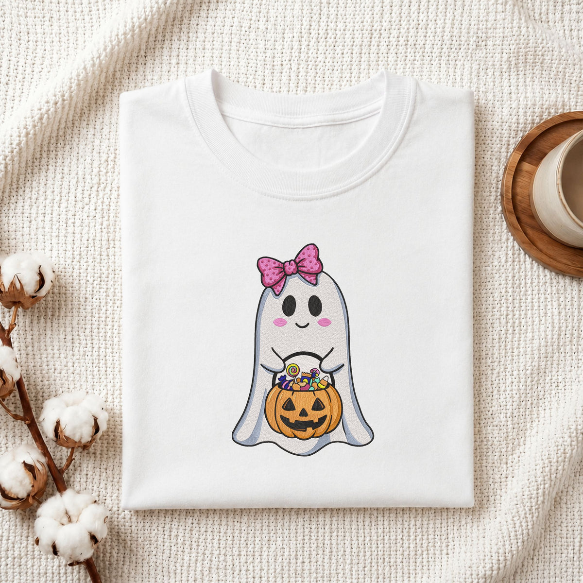 Folded white cotton t-shirt mockup featuring a cute ghost with a pink bow machine embroidery design, styled with cotton stems and a wooden cup
