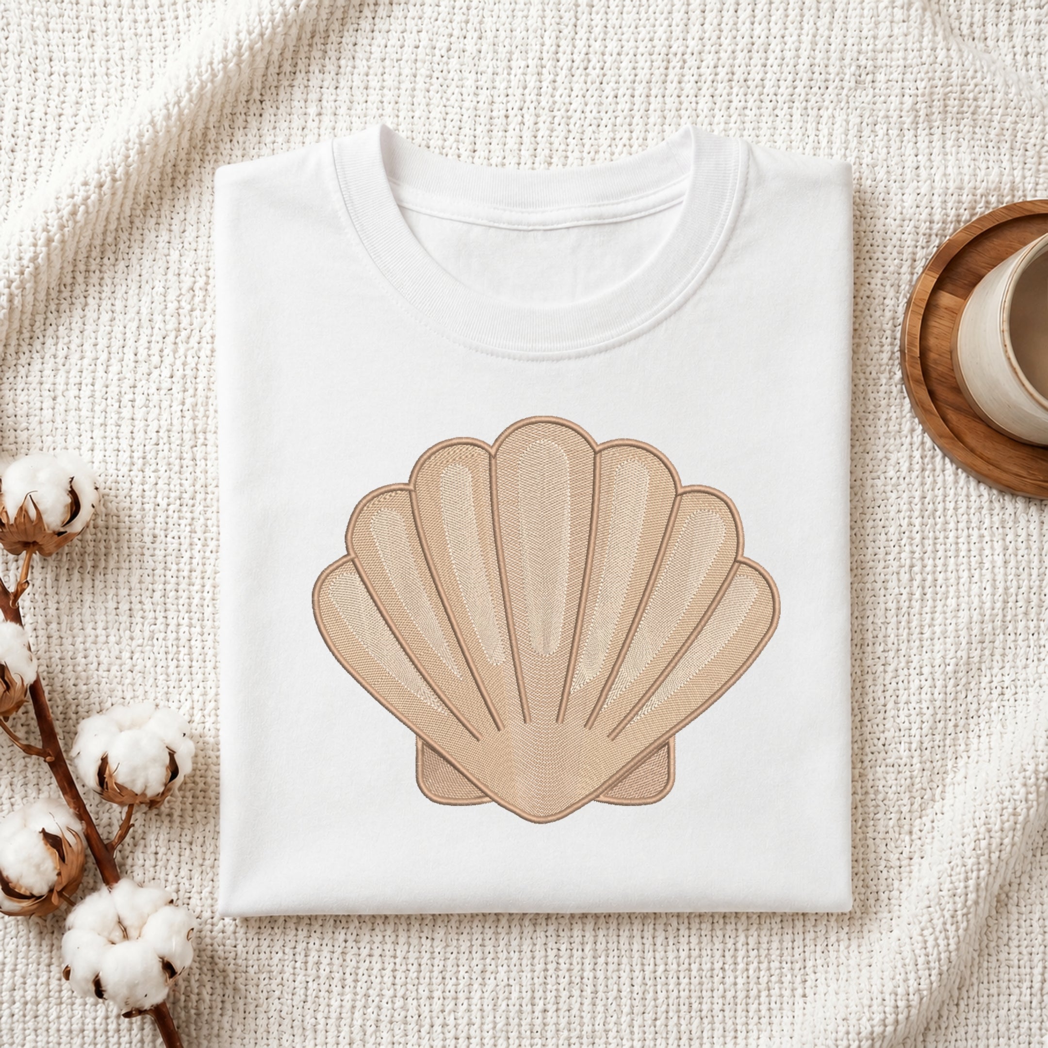 A folded white crewneck t-shirt featuring a large beige scallop shell graphic on the front, styled on a textured cream fabric with cotton bolls and a wooden cup.