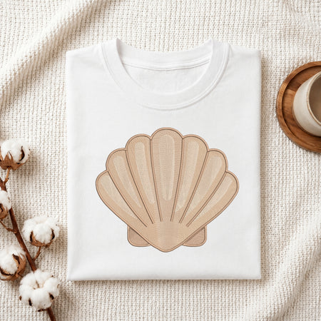 A folded white crewneck t-shirt featuring a large beige scallop shell graphic on the front, styled on a textured cream fabric with cotton bolls and a wooden cup.
