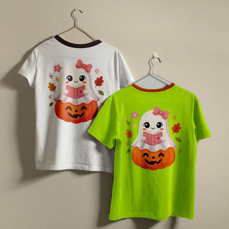 Two children's t-shirts hanging on metal hangers against a plain beige wall. The left shirt is white with a black collar trim and features a back graphic of a cute ghost reading a book over an orange pumpkin.
