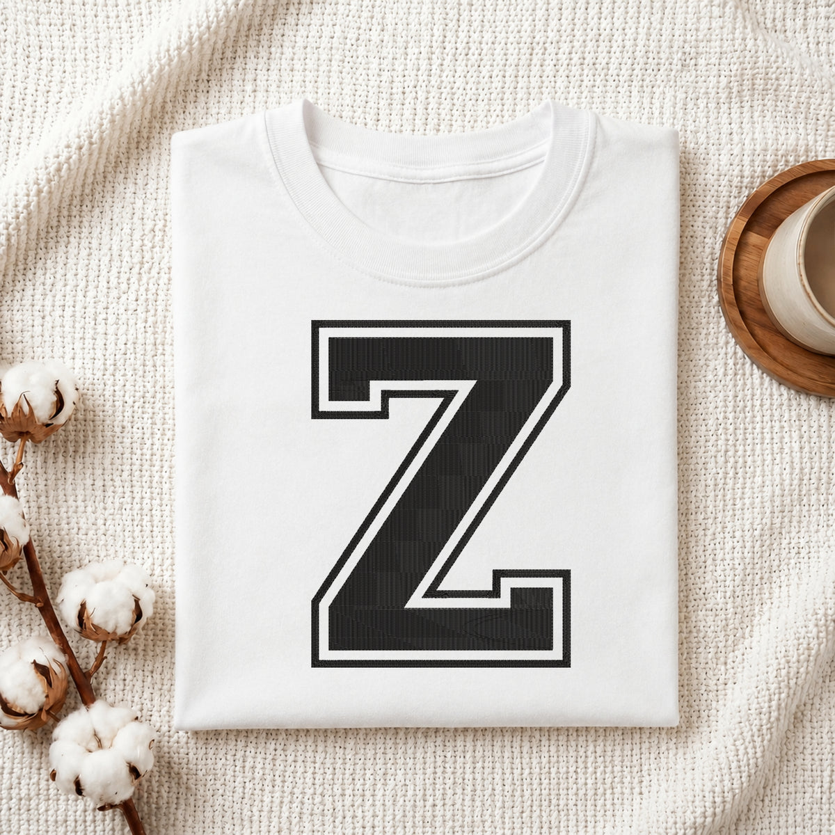 A folded white crewneck t-shirt with a large black block letter 'Z' outlined in white on the front, styled on a textured cream fabric with cotton bolls and a wooden cup.