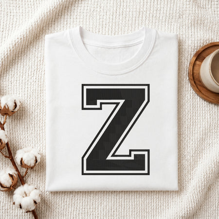 A folded white crewneck t-shirt with a large black block letter 'Z' outlined in white on the front, styled on a textured cream fabric with cotton bolls and a wooden cup.