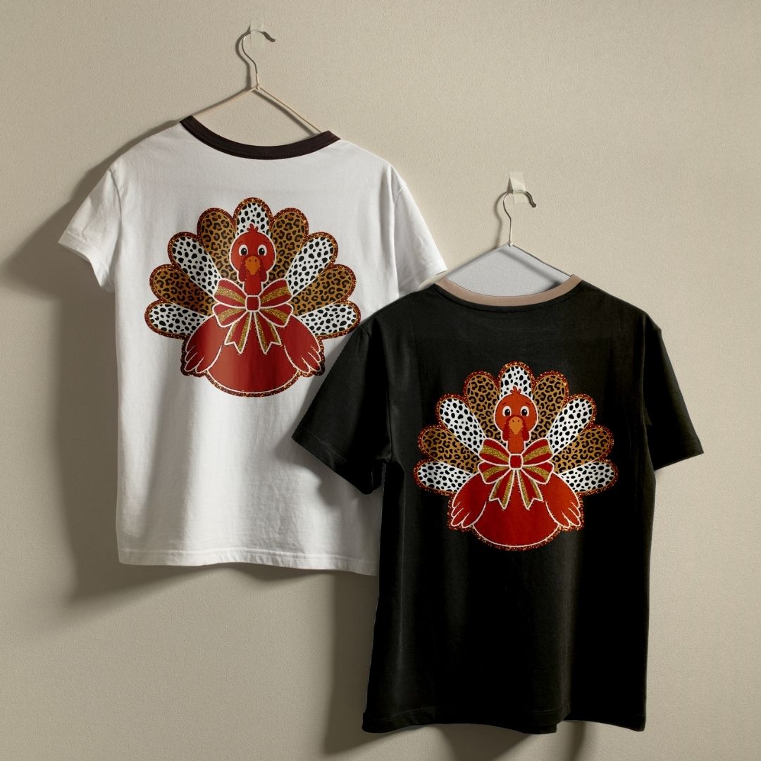 Two short-sleeve ringer T-shirts hanging on hangers against a plain beige wall, showing the front and back. The white shirt on the left has a dark brown collar and a large turkey graphic on the back with red, orange, brown, and black patterned feathers.