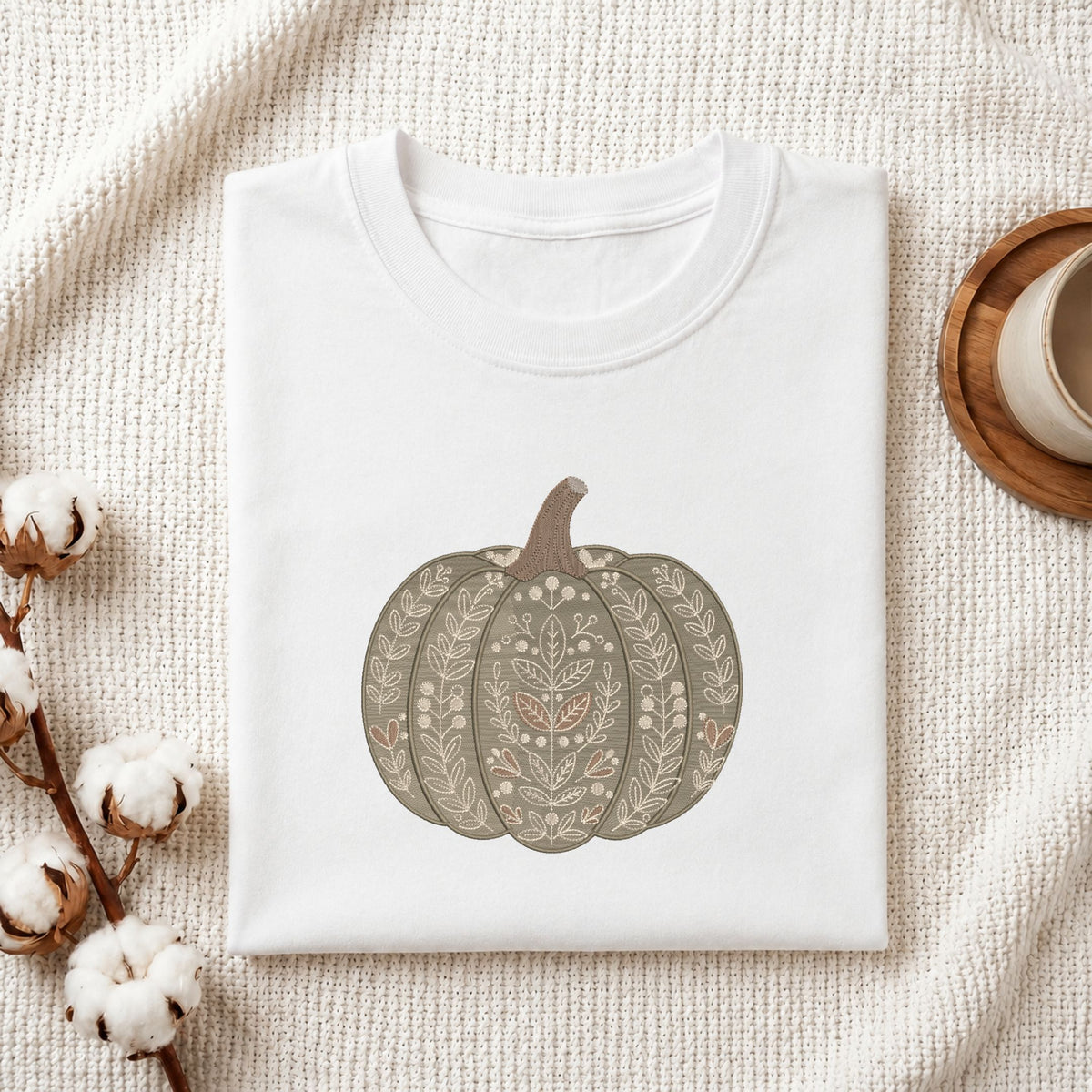 A folded white crewneck t-shirt featuring a decorative pumpkin graphic with intricate floral patterns, styled on a textured cream knit blanket with cotton bolls and a wooden cup in the background.