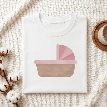 A folded white crewneck t-shirt with a pink and beige baby stroller graphic on the front, styled on a textured cream knit blanket with cotton bolls and a wooden cup in the background.