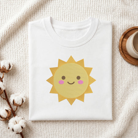 A folded white crewneck t-shirt featuring a cute yellow cartoon sun graphic with a smiling face and pink cheeks, styled on a textured cream knit blanket with cotton bolls and a wooden cup in the background.