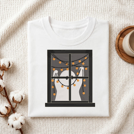 A folded white crewneck t-shirt displayed on a textured cream fabric background, decorated with cotton bolls and a cup on a coaster. The t-shirt features a graphic print of a window with ghostly white silhouettes and warm orange string lights.