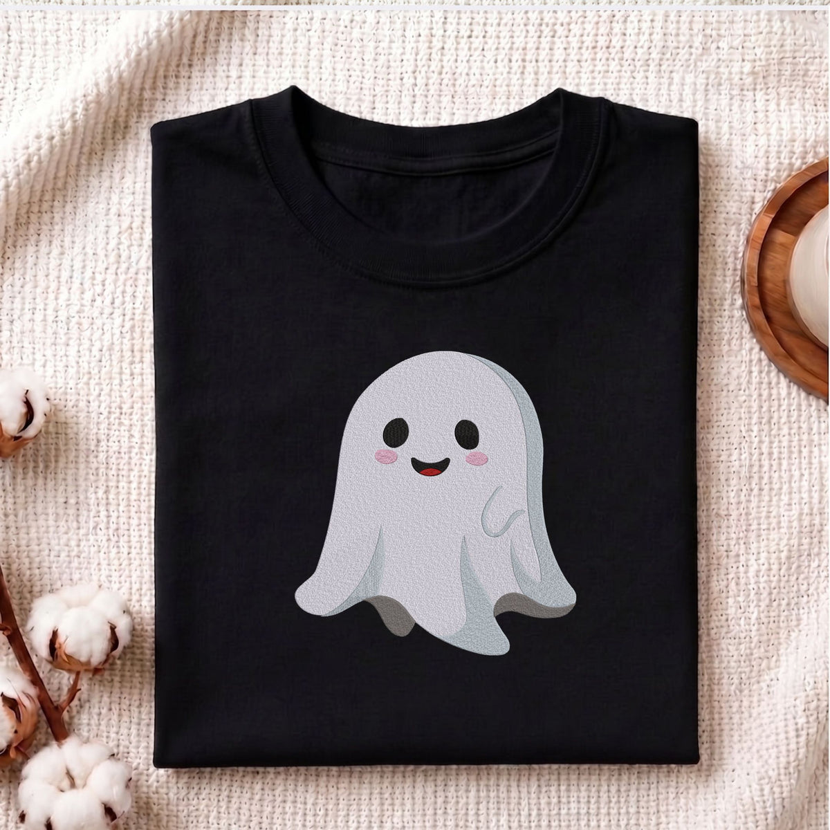 Folded black cotton t-shirt mockup featuring a cute smiling ghost machine embroidery design, styled with cotton stems and a wooden cup   