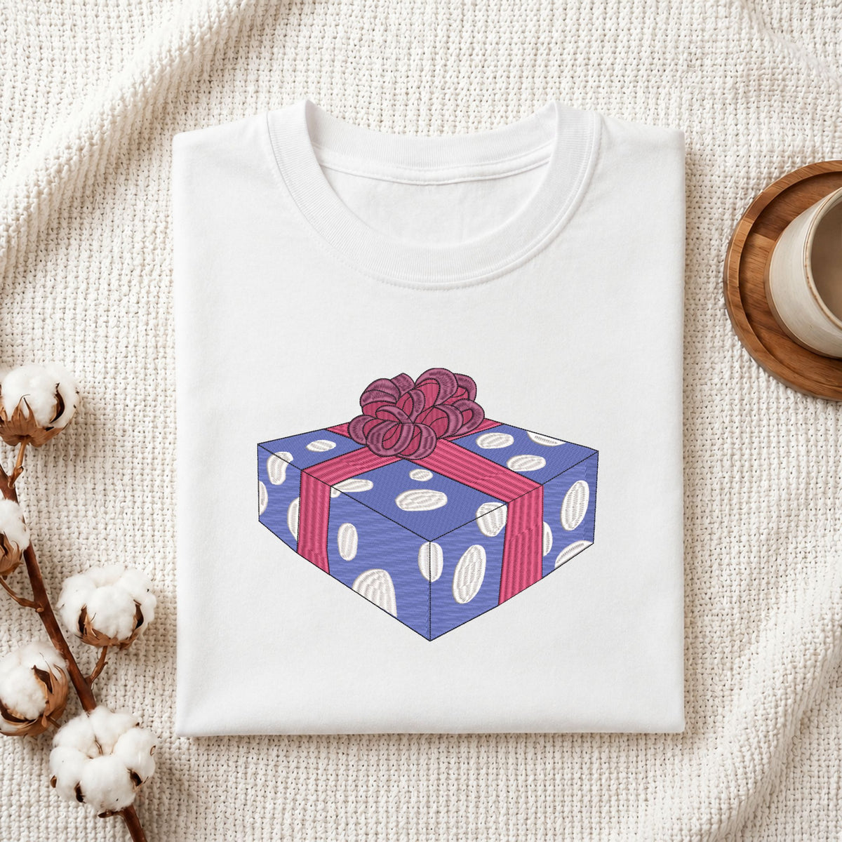 Folded white cotton t-shirt mockup featuring a wrapped gift box machine embroidery design, styled with cotton stems and a wooden cup   