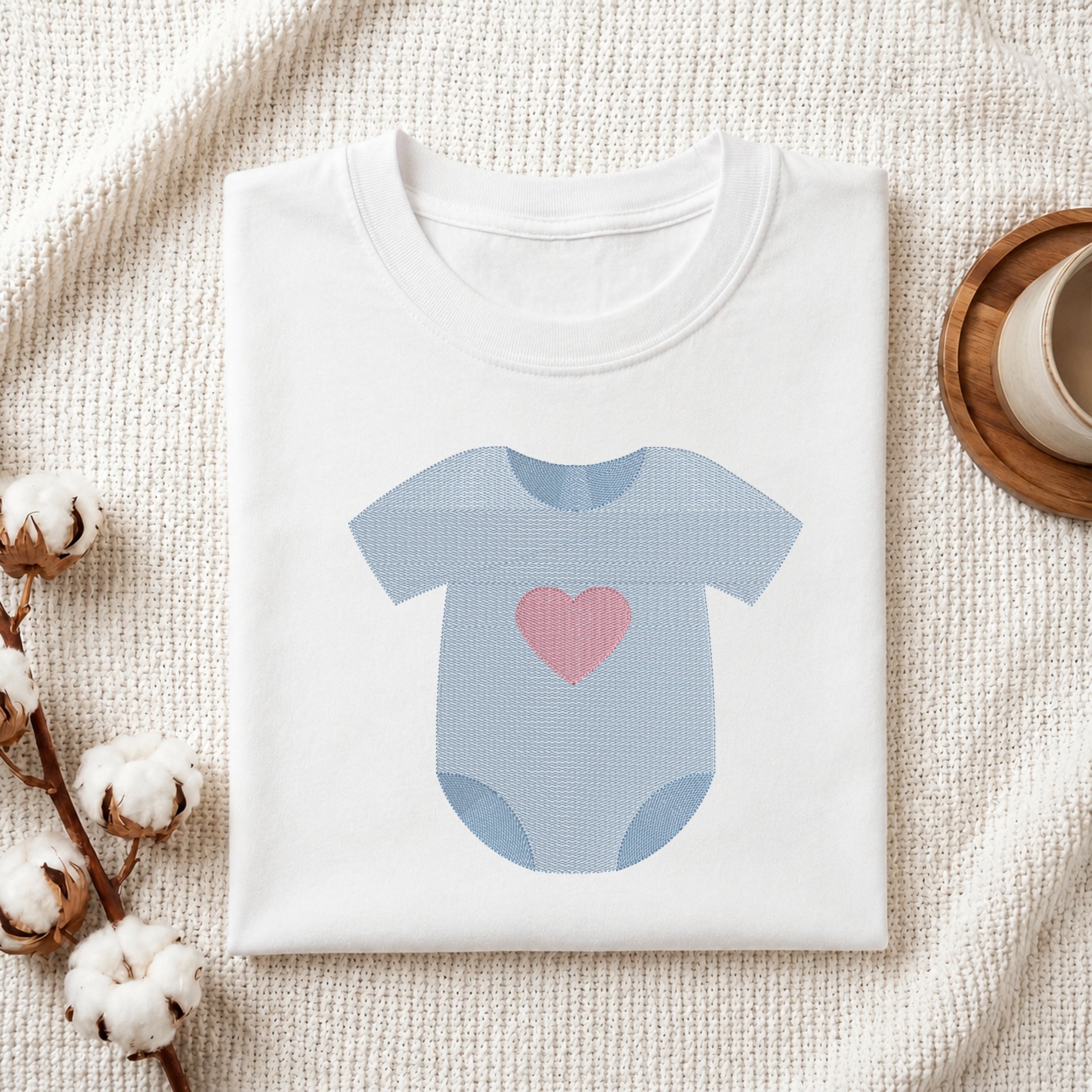 A folded white baby bodysuit or t-shirt with a light blue onesie graphic and a pink heart on the front, styled on a textured cream fabric with cotton stems and a small cup in the background.