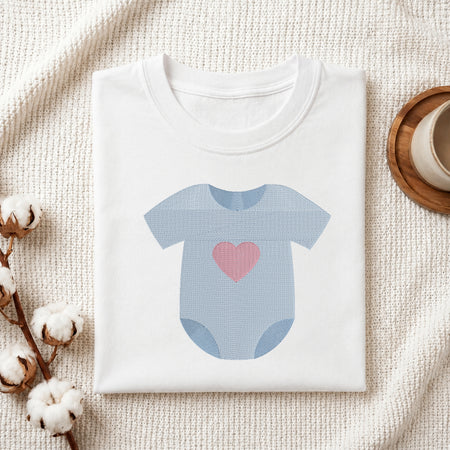 A folded white baby bodysuit or t-shirt with a light blue onesie graphic and a pink heart on the front, styled on a textured cream fabric with cotton stems and a small cup in the background.