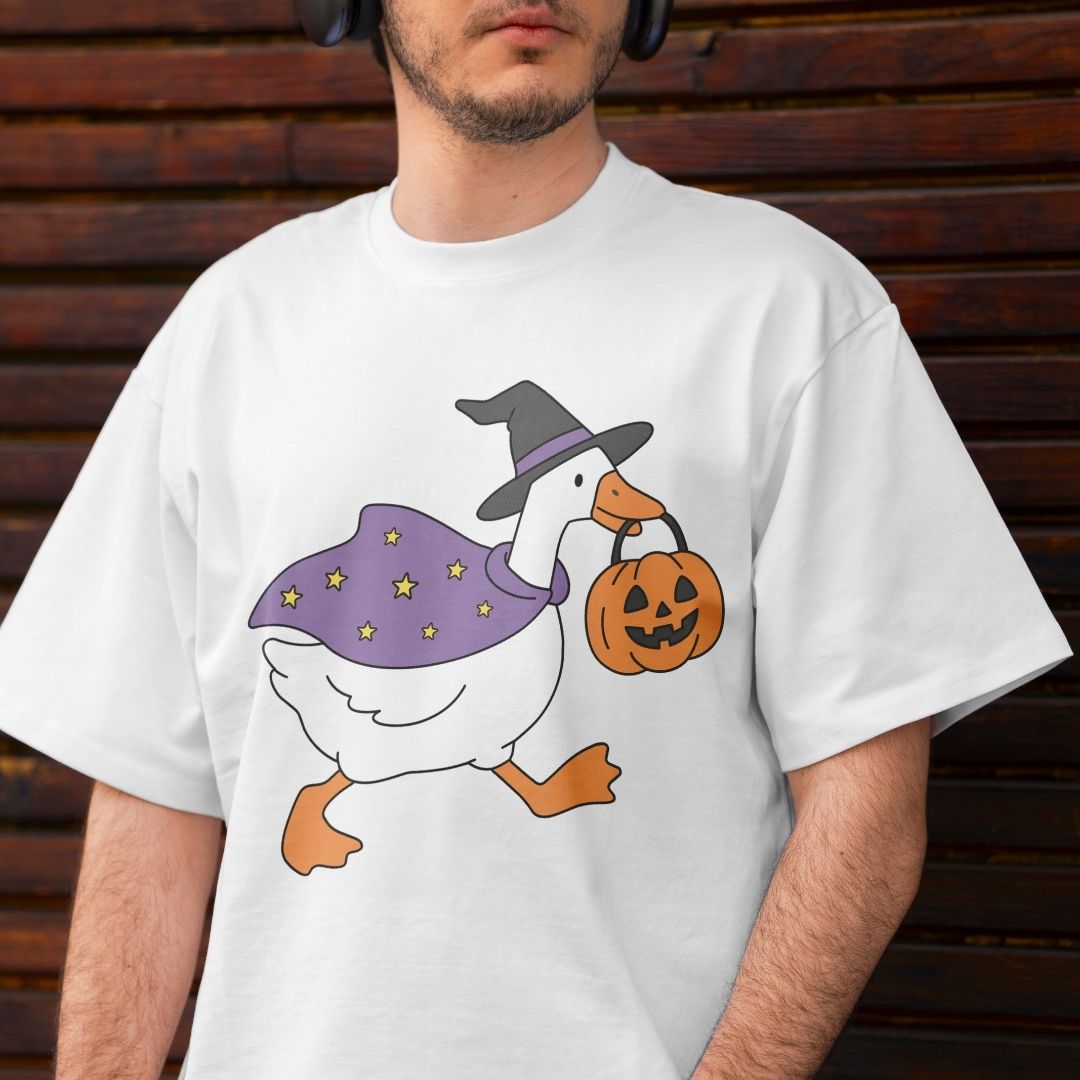 A man wearing a white crewneck t-shirt with a graphic of a white goose dressed in a purple witch cape and hat, carrying an orange jack-o'-lantern pumpkin bucket, set against a dark brown wooden background.