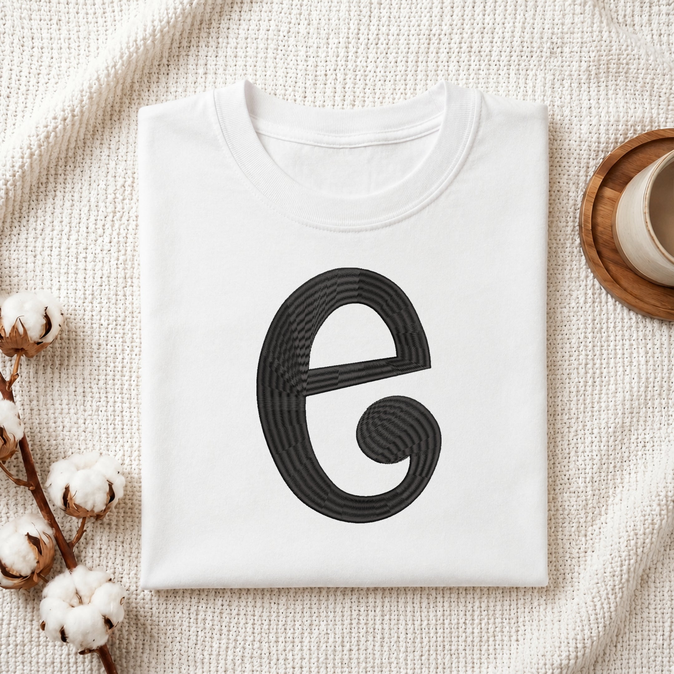 A folded white crewneck t-shirt with a large black stylized letter 'e' graphic on the front, styled on a textured cream knit blanket with cotton stems and a cup in the background.