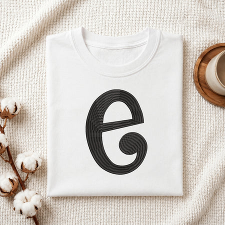 A folded white crewneck t-shirt with a large black stylized letter 'e' graphic on the front, styled on a textured cream knit blanket with cotton stems and a cup in the background.
