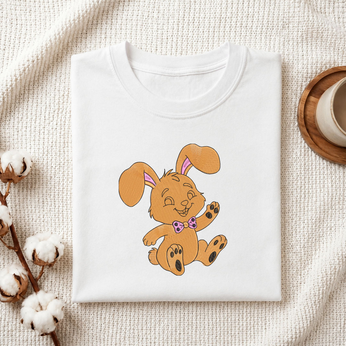 Folded white cotton t-shirt mockup featuring a cute golden-brown bunny machine embroidery design, styled with cotton stems and a wooden cup.