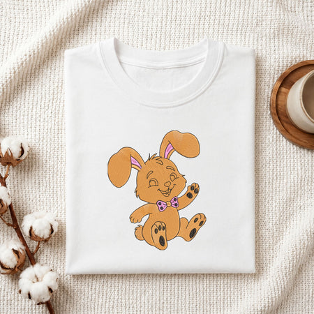 Folded white cotton t-shirt mockup featuring a cute golden-brown bunny machine embroidery design, styled with cotton stems and a wooden cup.
