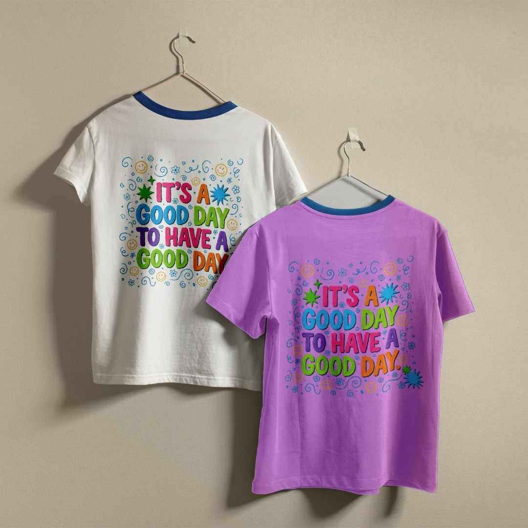 Two ringer-style short-sleeve T-shirts hanging on hangers against a plain beige wall. The left shirt is white with a blue collar and a colorful retro-style text graphic on the back reading 'IT'S A GOOD DAY TO HAVE A GOOD DAY.