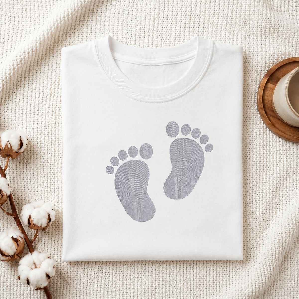 A folded white crewneck t-shirt with a gray baby footprint graphic on the front, styled on a textured cream knit blanket with cotton bolls and a wooden cup in the background.