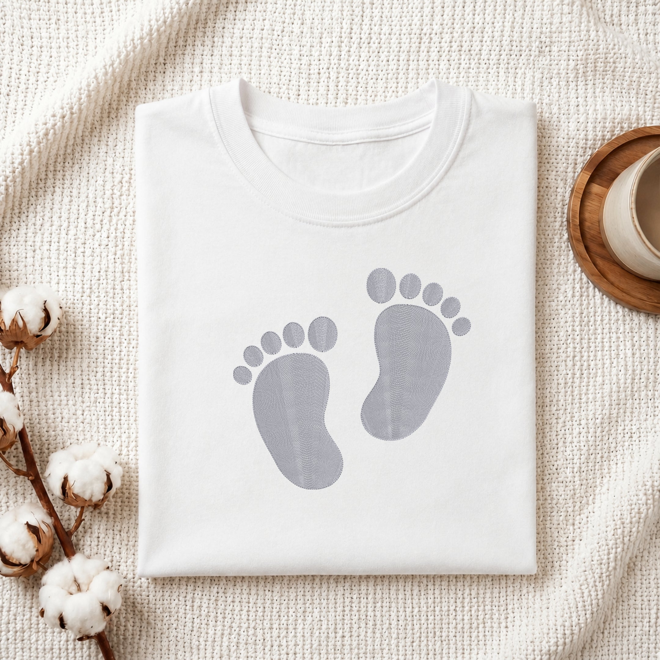 A folded white crewneck t-shirt with a gray baby footprint graphic on the front, styled on a textured cream knit blanket with cotton bolls and a wooden cup in the background.