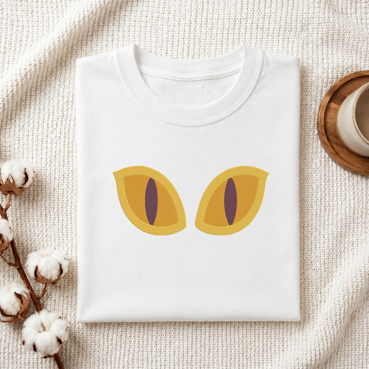A folded white t-shirt with a graphic of two yellow cat eyes, styled on a textured cream blanket with cotton bolls and a cup on a wooden tray.