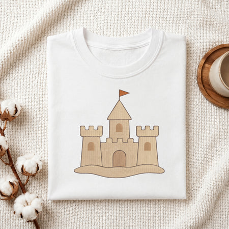 A folded white T-shirt with a cartoon sandcastle graphic on the front, styled on a textured cream fabric with cotton bolls and a wooden cup in the background.
