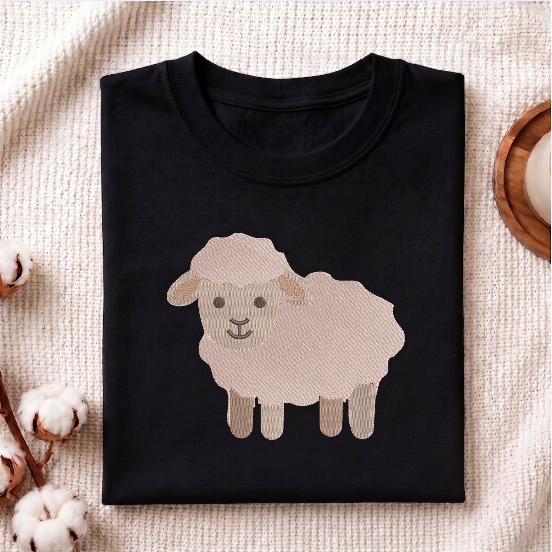 A folded black crewneck t-shirt featuring a cartoon sheep graphic on the front, styled on a textured cream fabric with cotton bolls and a wooden coaster.