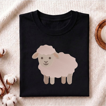 A folded black crewneck t-shirt featuring a cartoon sheep graphic on the front, styled on a textured cream fabric with cotton bolls and a wooden coaster.