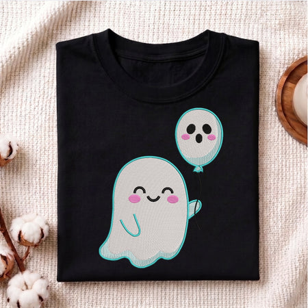 A folded black short-sleeve T-shirt with a cute cartoon ghost and balloon graphic, styled on a textured cream fabric with cotton bolls and a wooden slice prop.